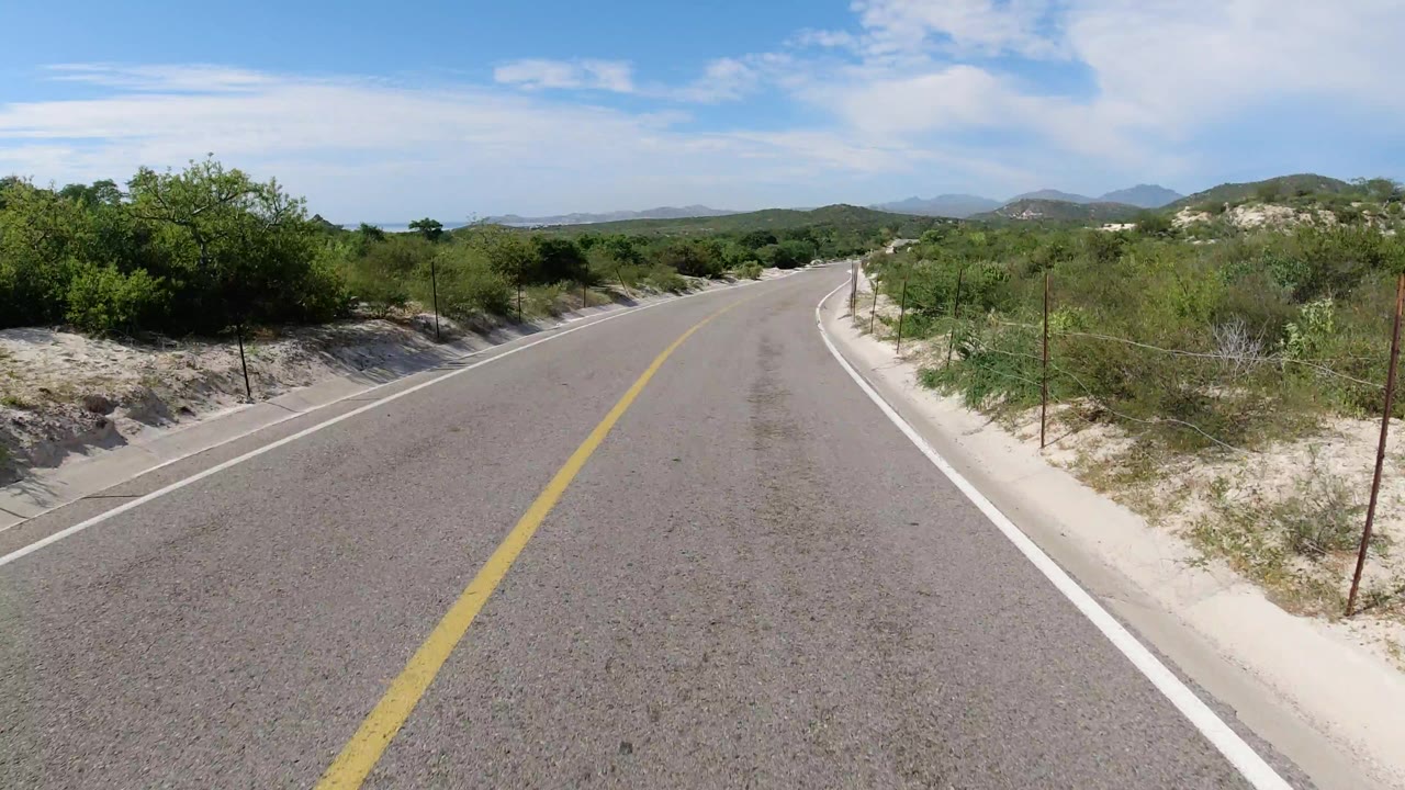 Forward road view in Los Cabos, Mexico, showing the yellow line near center, white shoulder markings, scrubby vegetation on both sides, low hills ahead, and no visible people.