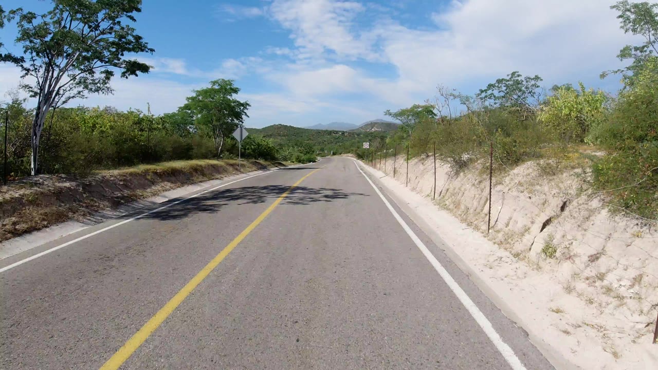 Start of the Los Cabos road clip, looking down an empty two-lane asphalt road with sandy banks, green brush, a blue sky, and no visible people.