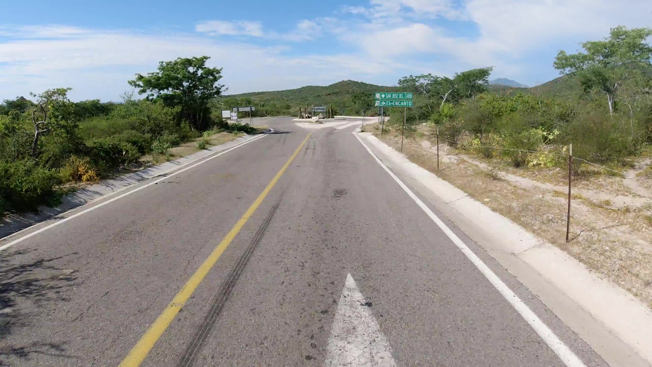Forward-facing view of a paved road in Los Cabos, Mexico, with a yellow center line, roadside brush, distant hills, blue sky, and no visible people.