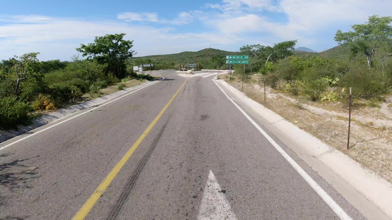 The Los Cabos road remains centered as it approaches a junction area, with scrubby trees on both sides, pale shoulders, distant hills, and no visible people.
