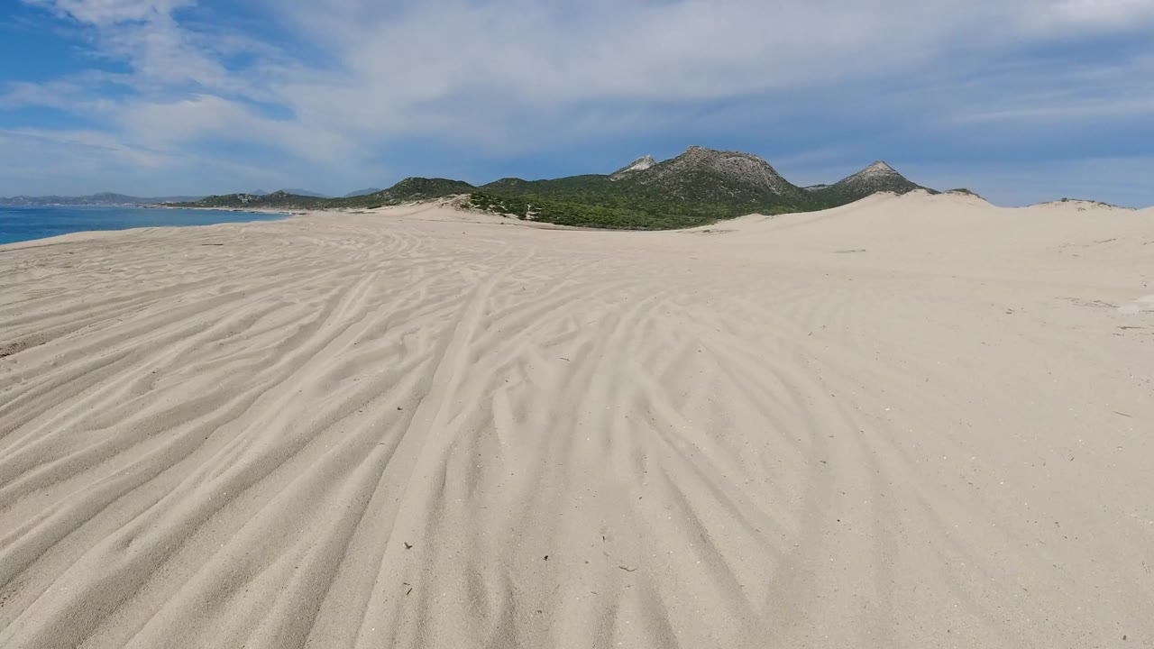 Wide view of rippled sand in Los Cabos, Mexico, with blue water at the left edge, green coastal hills in the background, and no visible people.