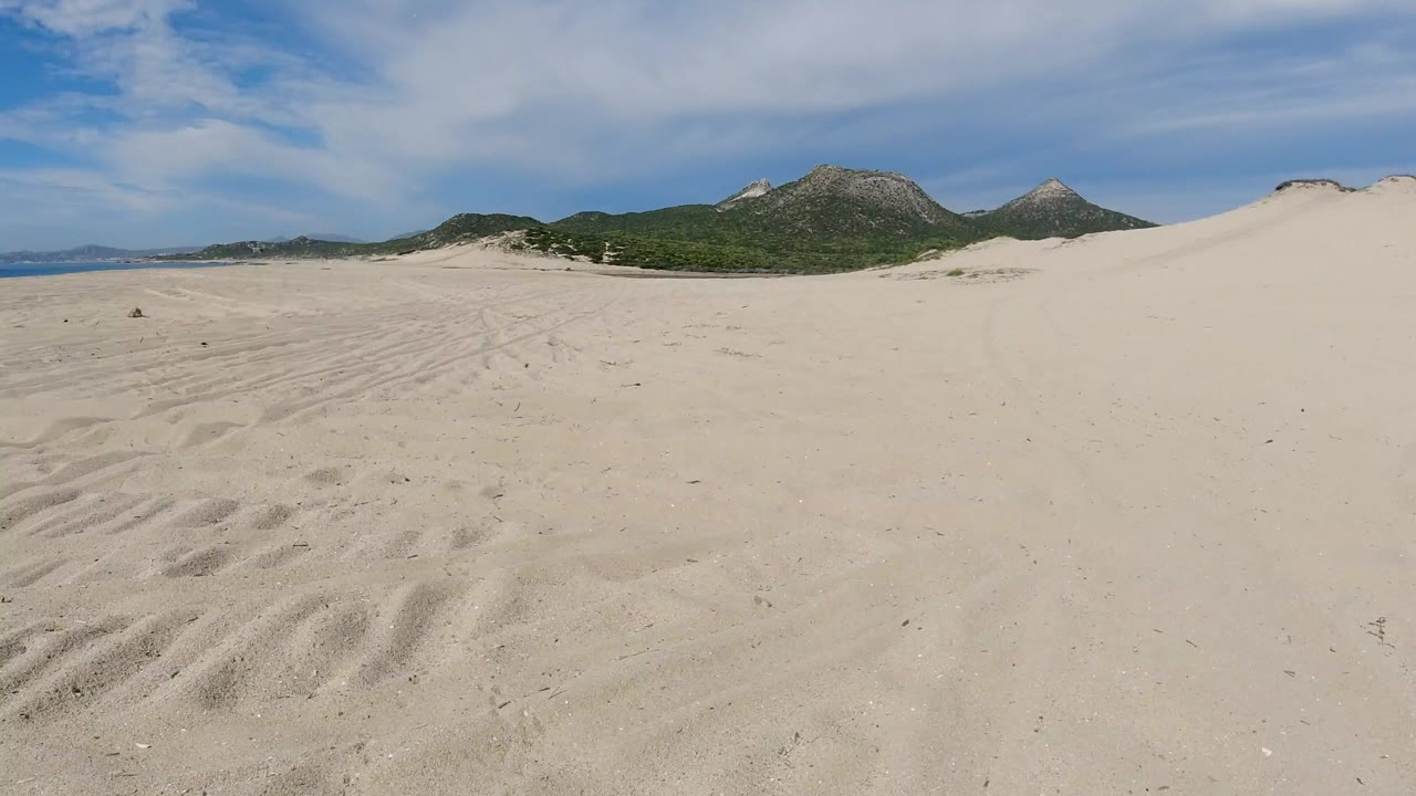 Wide view of a sandy beach near Los Cabos with pale foreground tracks, low green hills, rocky peaks, and no visible people.