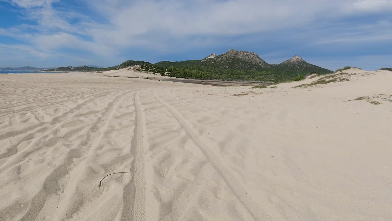 Opening frame of a wide sandy Los Cabos landscape with tire tracks across pale foreground sand, low dunes, distant rocky mountains, thin cloud, and no visible people.