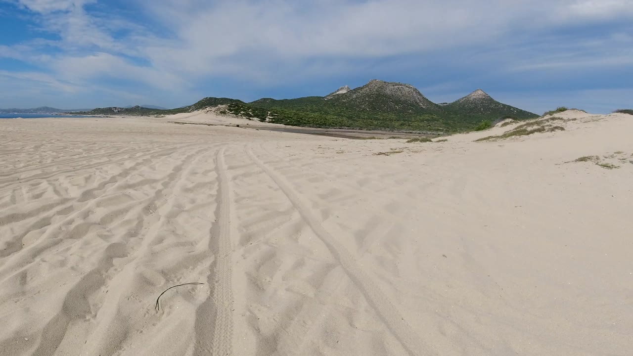 Second frame shows parallel tire marks and rippled sand leading toward green scrub and rocky mountains under a blue, lightly clouded Los Cabos sky, with no visible people.