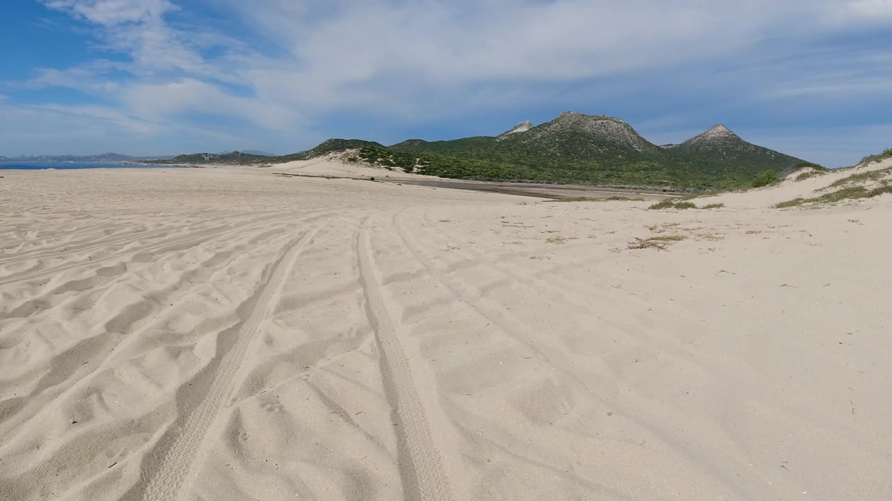 Empty pale sand with tire ruts, scrubby dunes, mountains, and clouded sky.