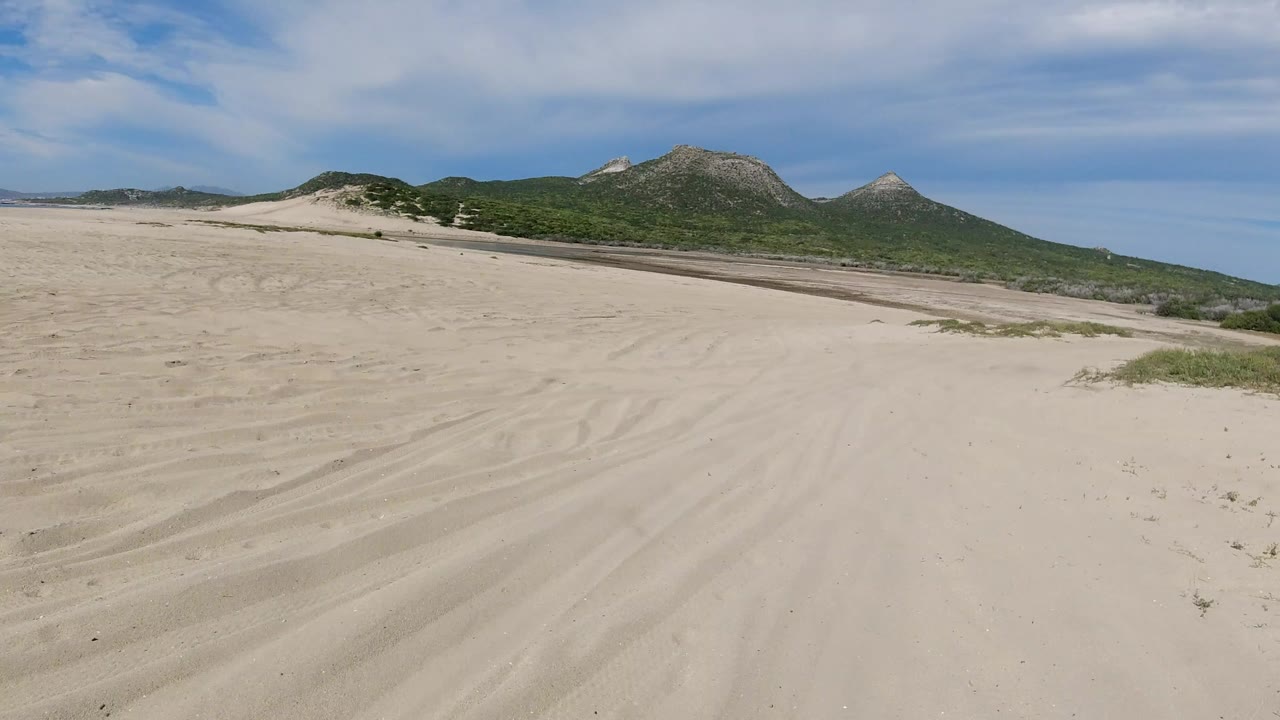 Aerial-looking view of a sandy track in Los Cabos, Mexico, with tire marks in pale sand, scrub-covered hills, blue sky, and no visible people.