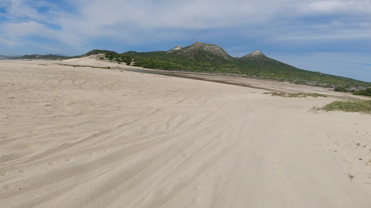 The sandy route continues across open Los Cabos terrain, with faint tire lines, low brush, green hills, and no visible people.