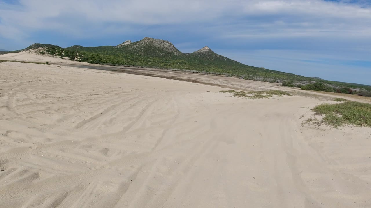 Broad pale sand foreground with tracks, scrub, and hills across the horizon.