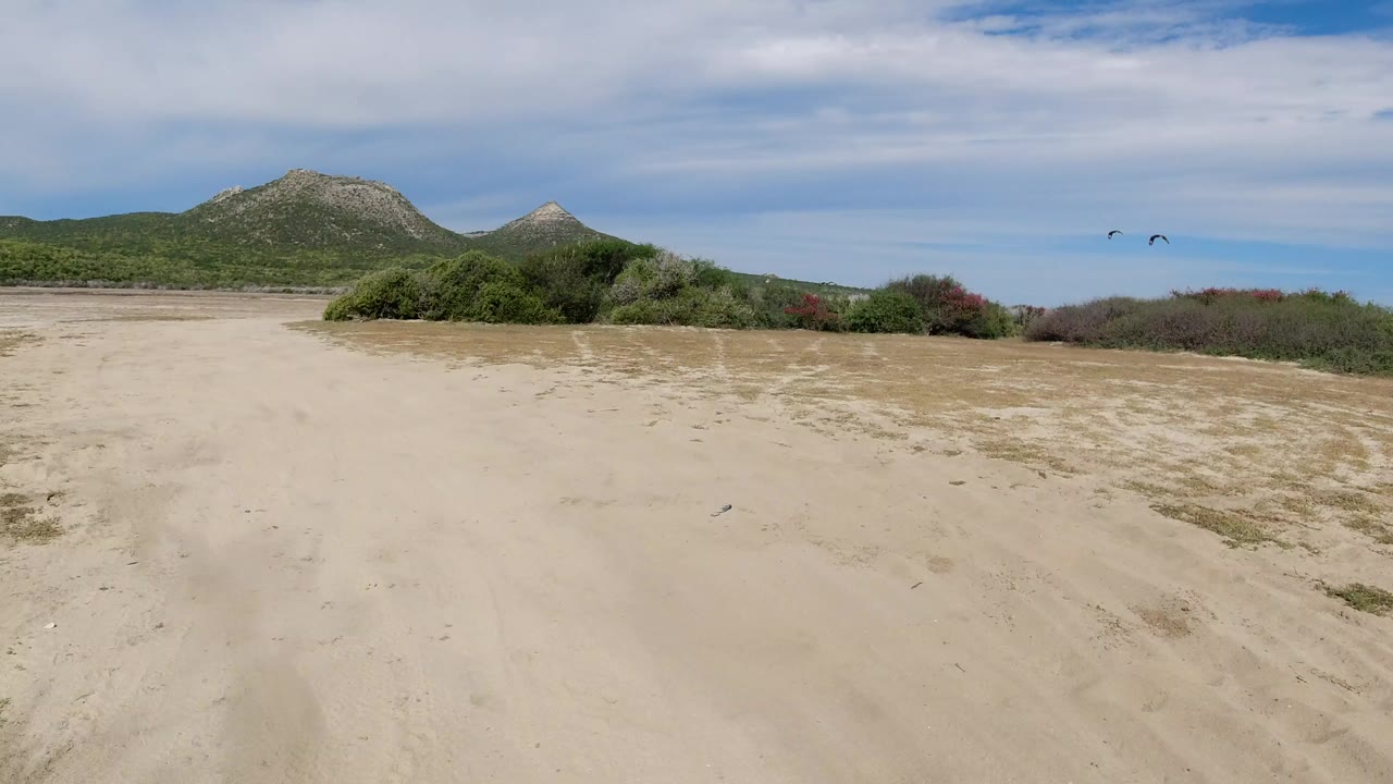 Sandy dirt track with scrub bushes and mountains under a cloudy blue sky.