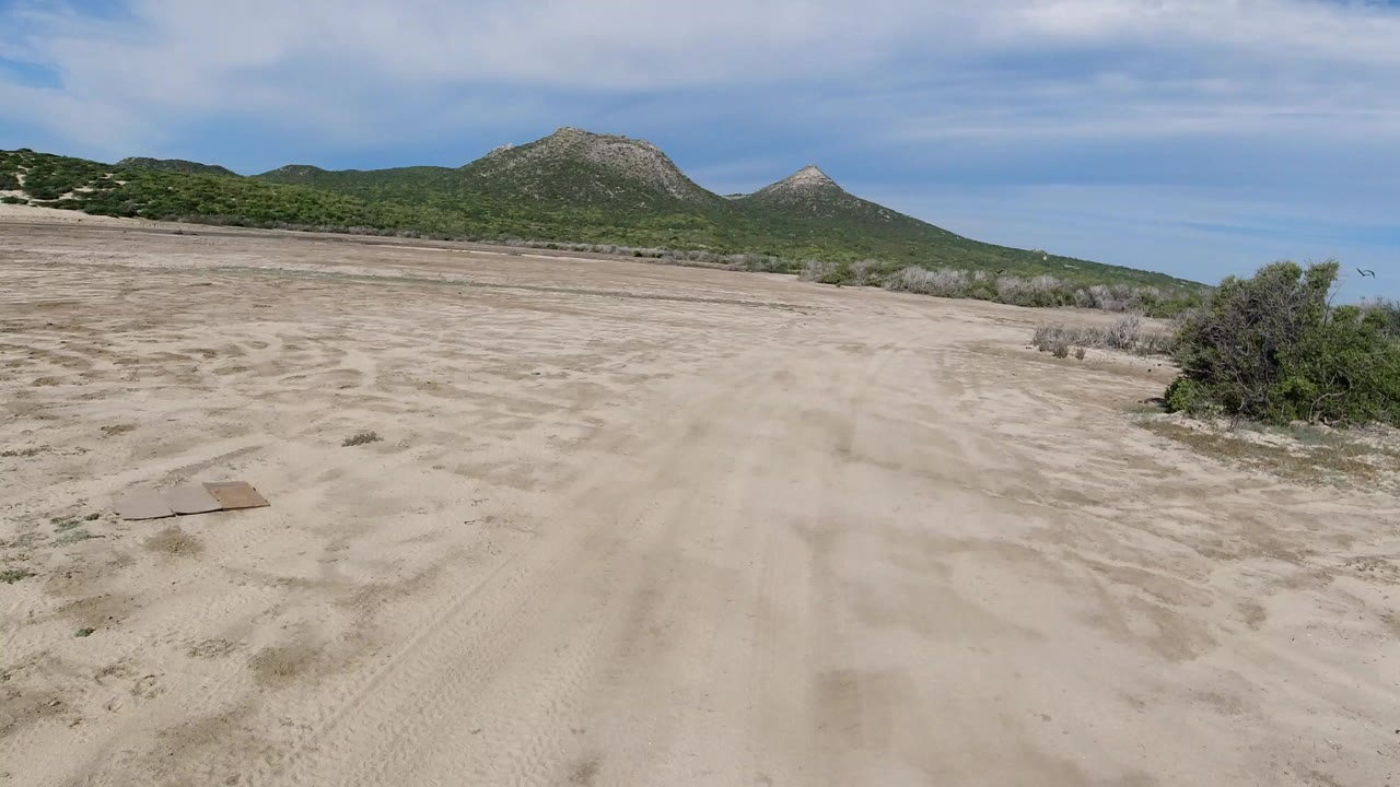 Wide aerial view in Los Cabos, Mexico, with a sandy track in the foreground, scrub along the edge, green hills in the distance, and no visible people.