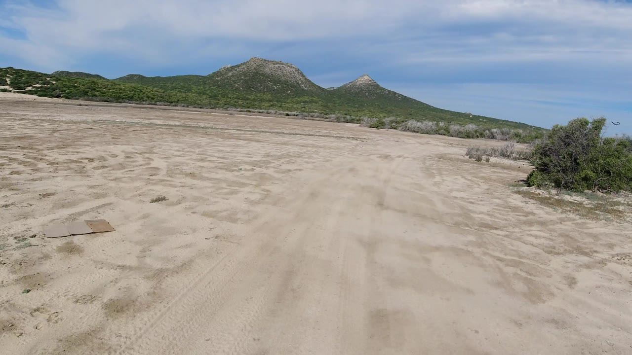 Aerial frame in Los Cabos, Mexico, looking over pale sandy ground toward scrubland and two rounded mountains beneath streaked cloud cover, with no visible people.