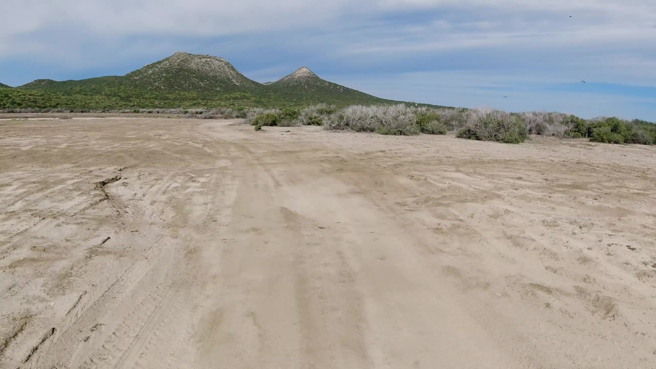 Wide view of a pale dirt road in Los Cabos, Mexico, with scrub brush, green hills, rocky peaks, thin clouds, and no visible people.