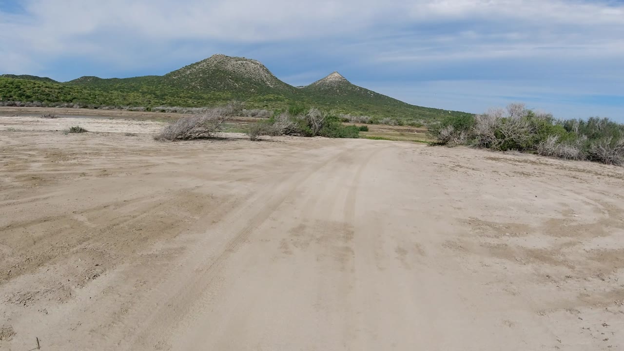 Dry sandy clearing with green hills and shrubs along the horizon.