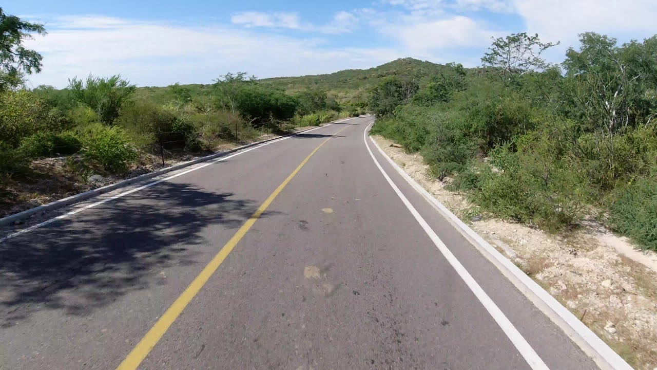 Forward windshield-style view of a paved road in Los Cabos, Mexico, with a yellow center line, white shoulders, green roadside brush, distant low hills, and no visible people.