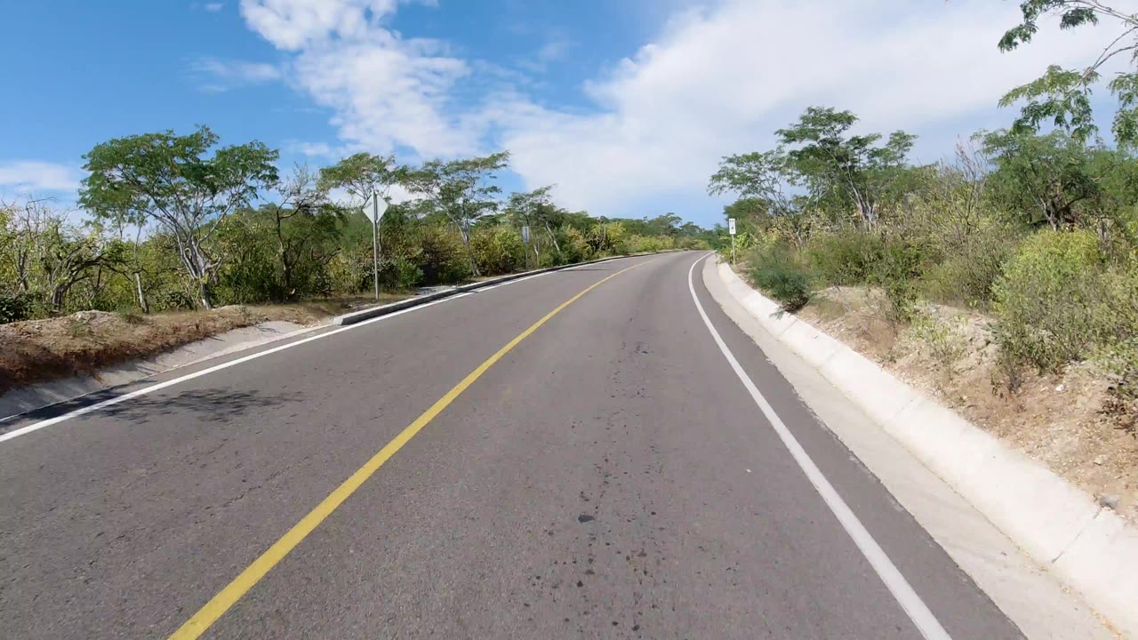 Start of a Los Cabos road-driving view with an empty paved lane, yellow centerline, white shoulder stripe, roadside scrub, trees, puffy clouds, and no visible people.