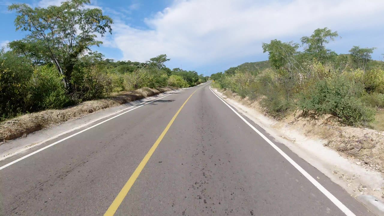 Forward road view in Los Cabos, Mexico, with a yellow centerline, white shoulders, green brush, low hills, blue sky, scattered cloud, and no visible people.