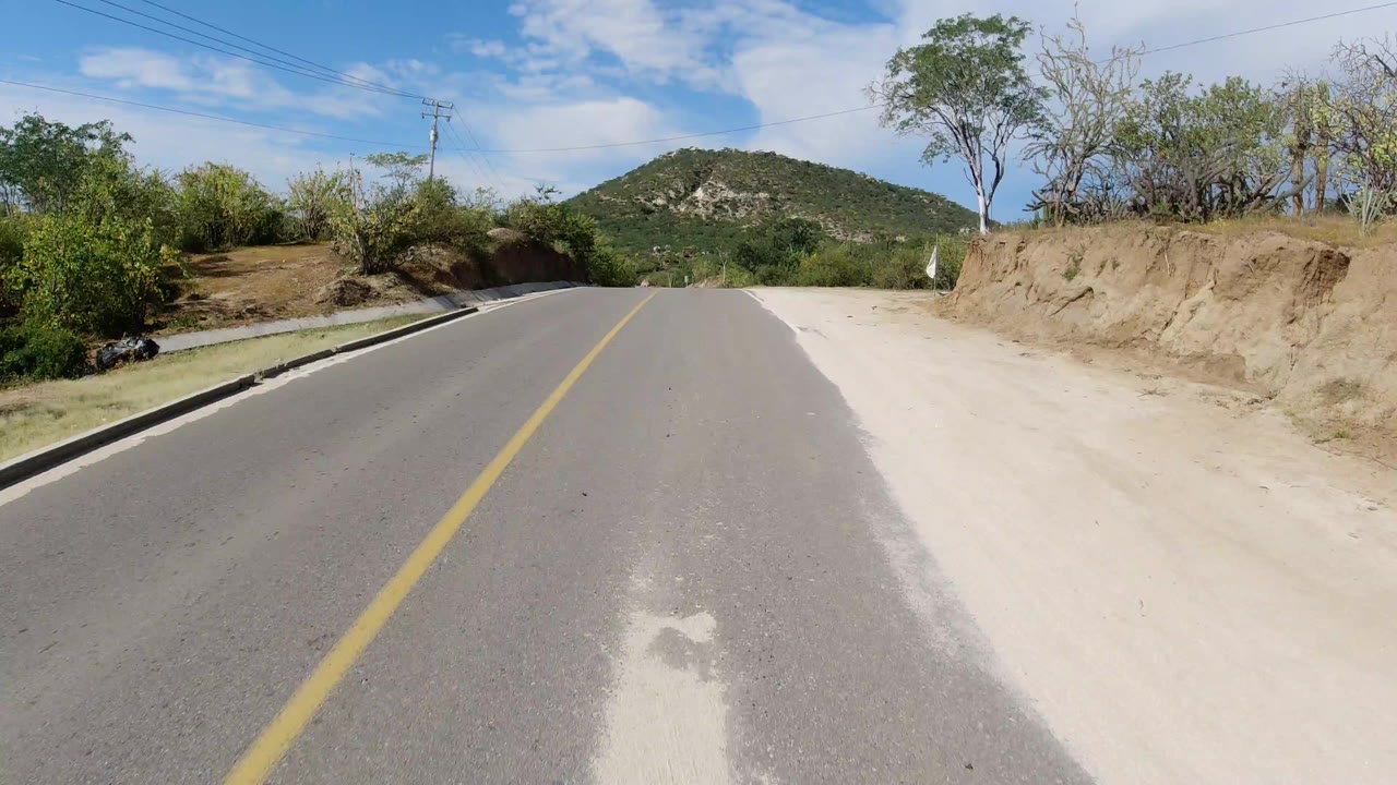 Los Cabos road view from a moving vehicle, with a yellow center line, sandy shoulder, scrub vegetation, utility lines, a low hill ahead, and no visible people.