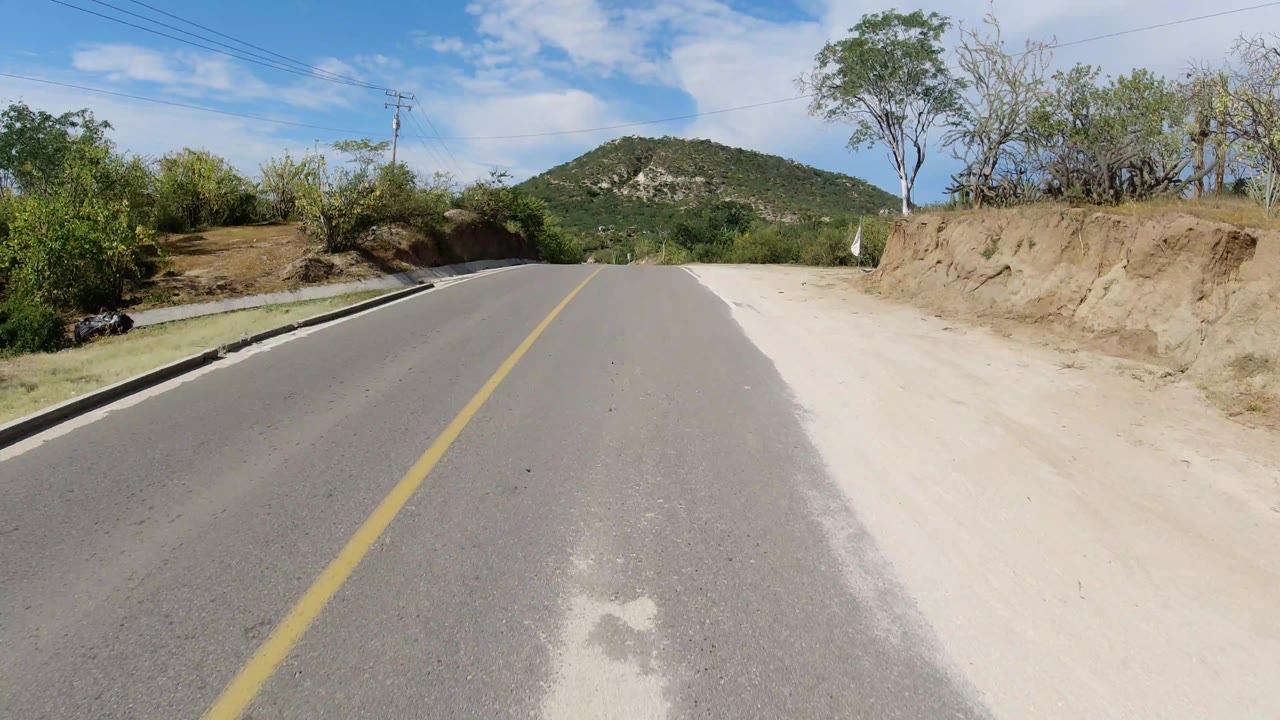 Forward road view in Los Cabos with asphalt foreground, pale dirt shoulders, scrubby plants on both sides, a rounded hill ahead, and no visible people.
