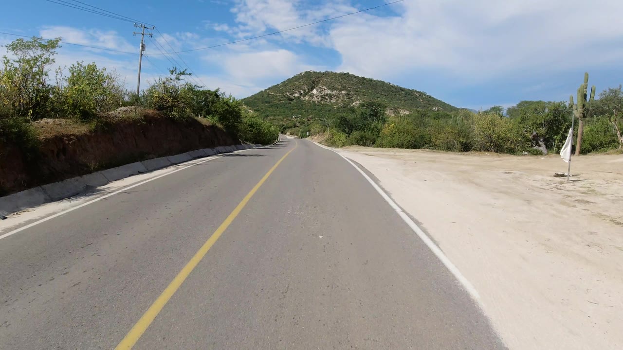 Empty paved road bordered by dirt shoulder, shrubs, cactus, and a hill under clouds.
