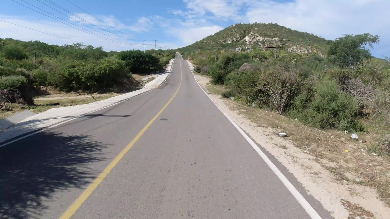 An empty paved two-way road in Los Cabos, Mexico runs toward a green rocky hill, with scrub bushes, pale shoulders, utility lines, bright daylight, and no visible people.