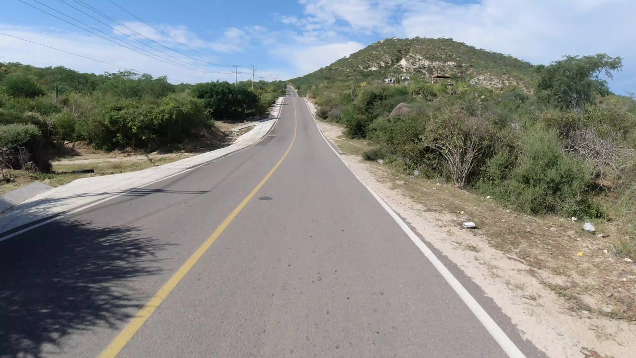 A forward view along the Los Cabos road shows the yellow center line, concrete drainage edges, dense roadside shrubs, a rocky hill ahead, overhead wires, and no visible people.