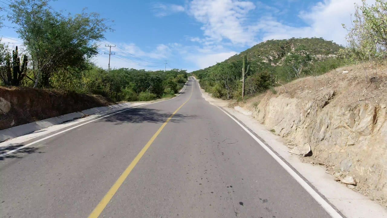 Forward road view in Los Cabos, Mexico, with a yellow center line leading toward a low rocky hill, desert shrubs, cacti, overhead wires, and no visible people.