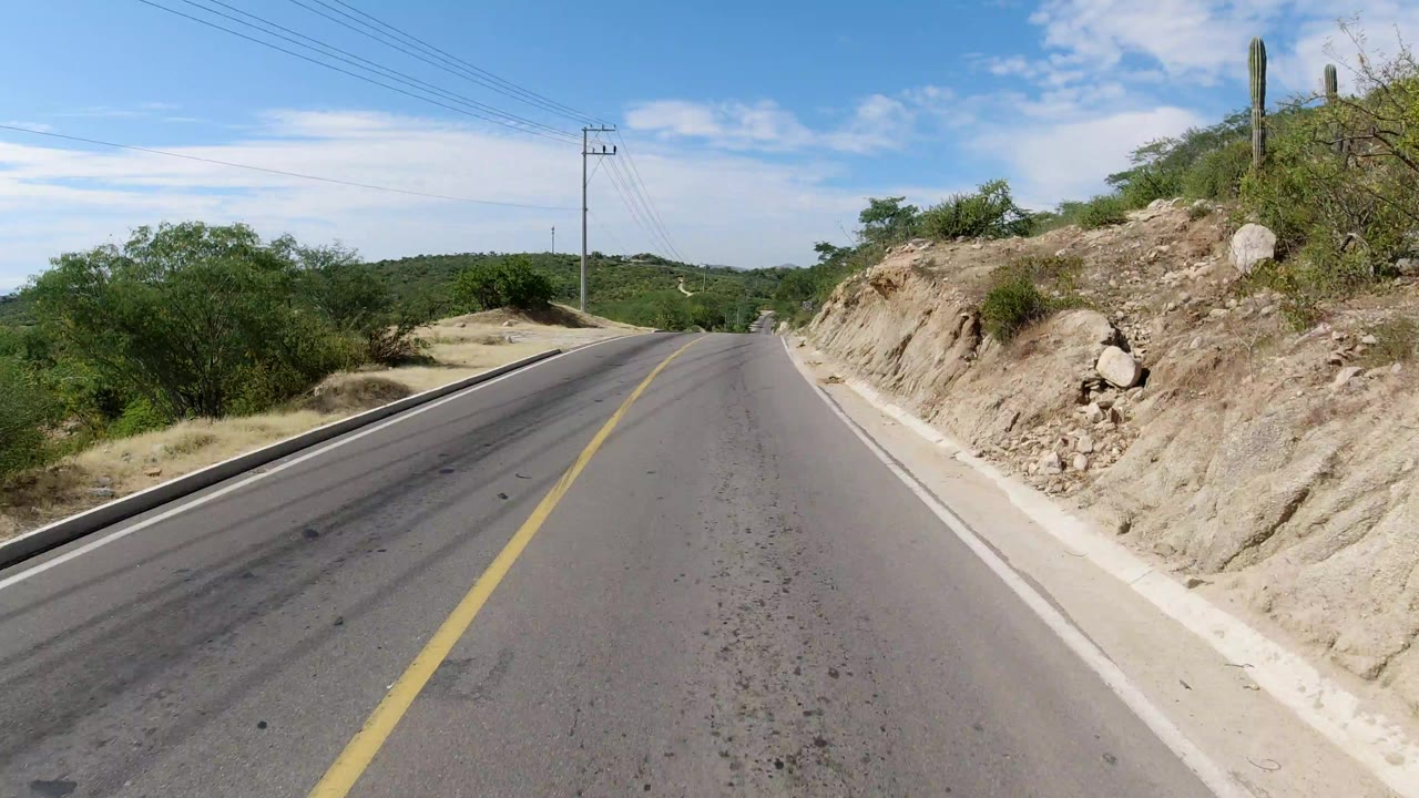 Early frame of a paved Los Cabos road climbing toward a low crest, with rocky shoulder, green brush, overhead utility wires, blue sky, and no visible people.
