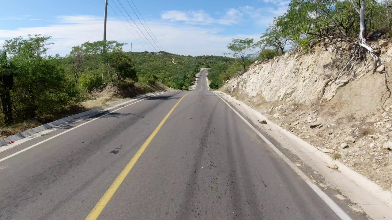 Forward view down an empty two-lane highway in Los Cabos, Mexico, with a yellow center line, pale rock bank, green brush, powerlines, and no visible people.
