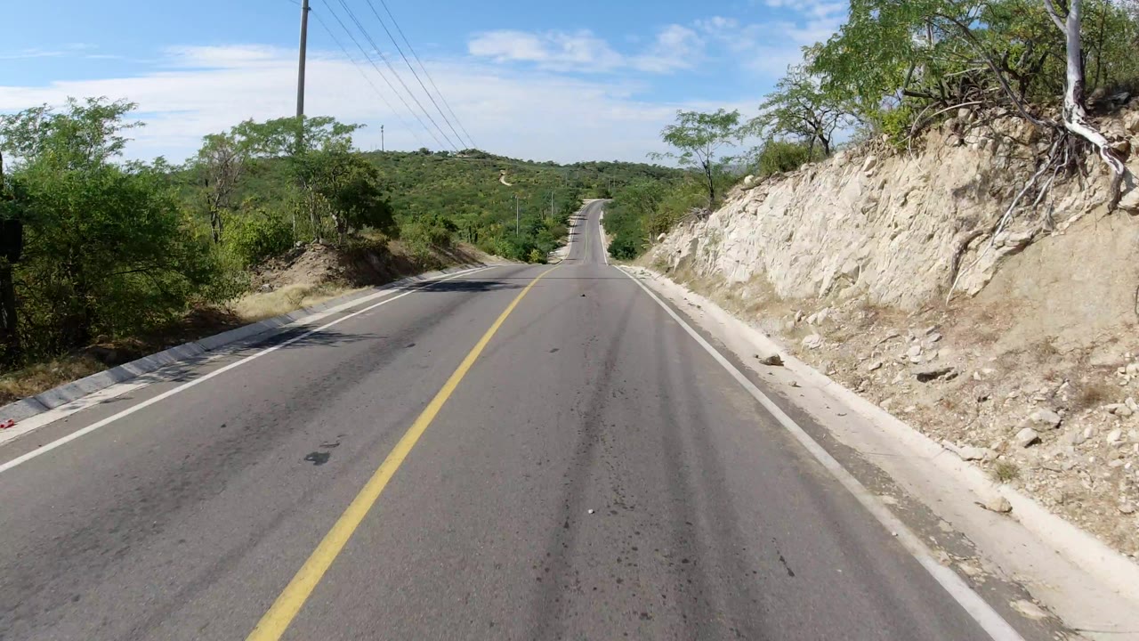 The highway remains centered in Los Cabos, Mexico, with rocky shoulder on the right, dense green brush to both sides, powerlines above, and no visible people.