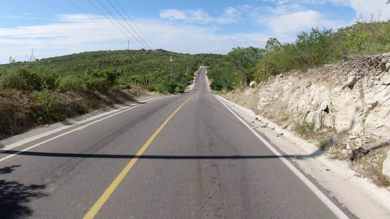 Straight empty highway bordered by rocky slope, brush, and low hills
