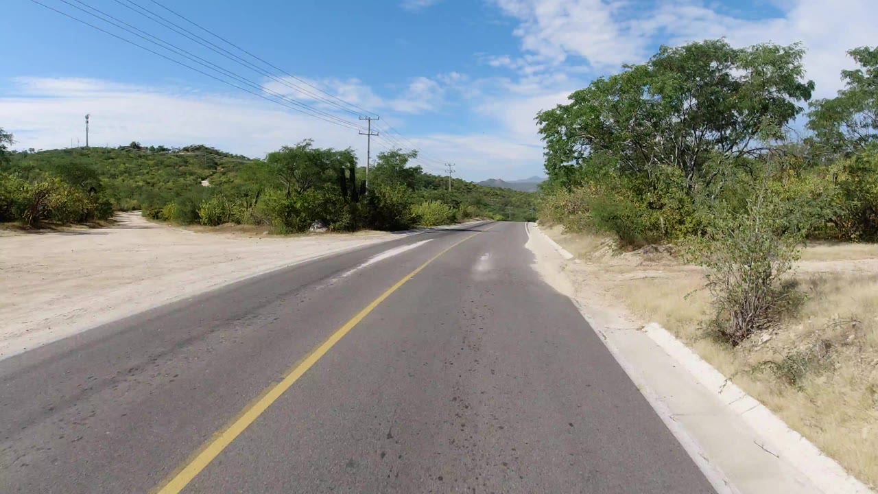 Opening road-level view in Los Cabos, Mexico, showing a two-lane paved road, sandy side track, green roadside brush, utility lines, low hills, and no visible people.