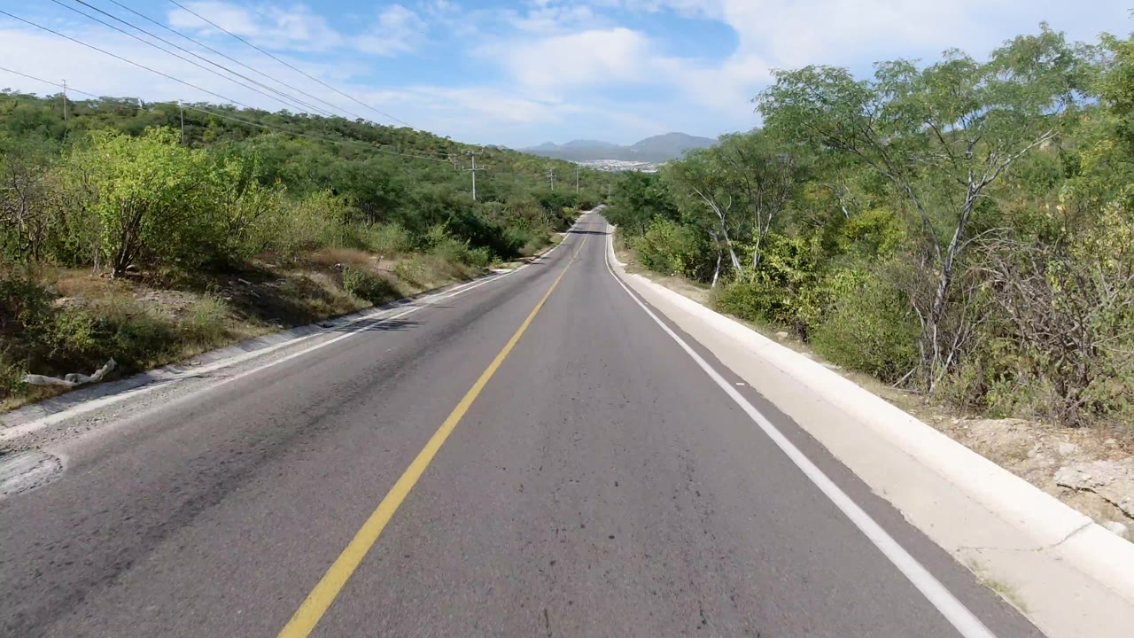 The road continues straight through Los Cabos, Mexico, with green brush close to the shoulders, utility lines overhead, distant hills, and no visible people.