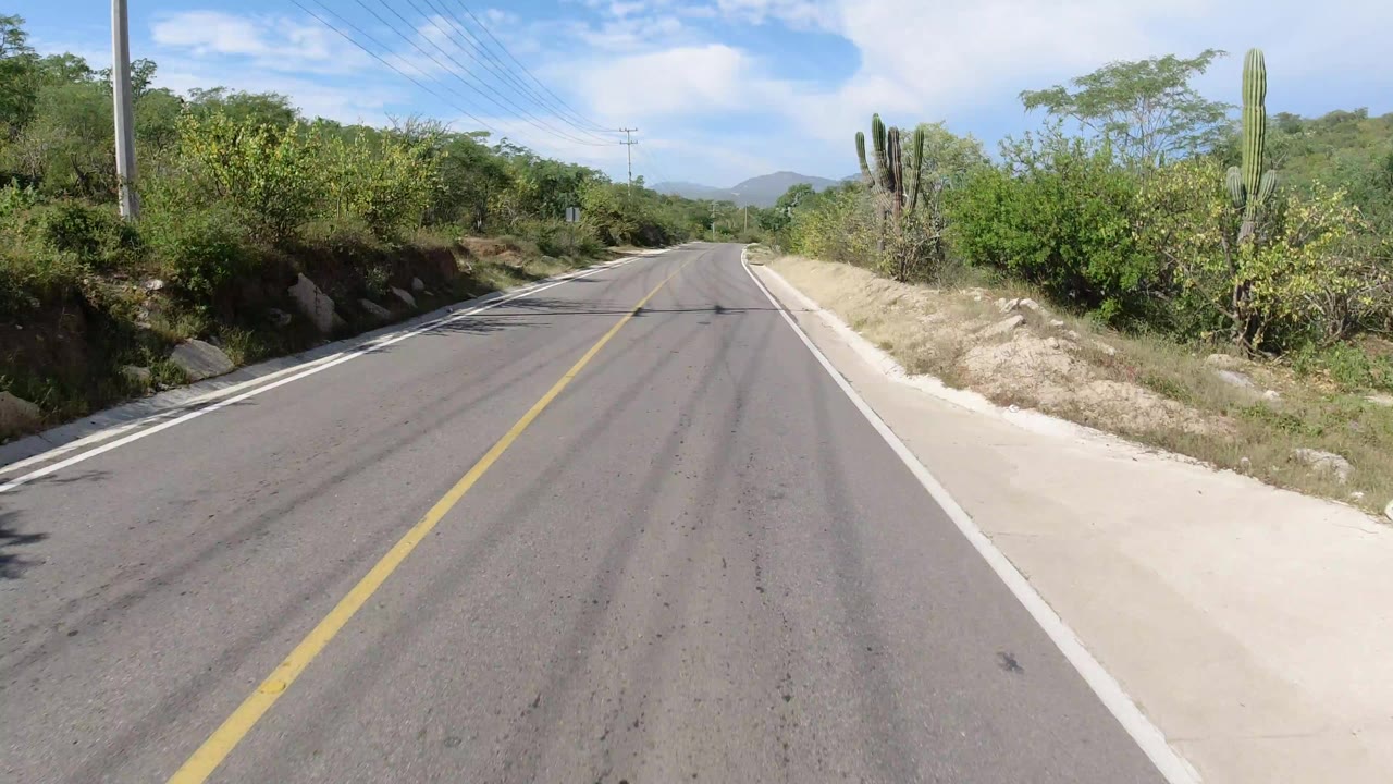Opening view of an empty two-lane road in Los Cabos, Mexico, with a yellow centerline, roadside shrubs, cactus, utility poles, distant hills, and no visible people.