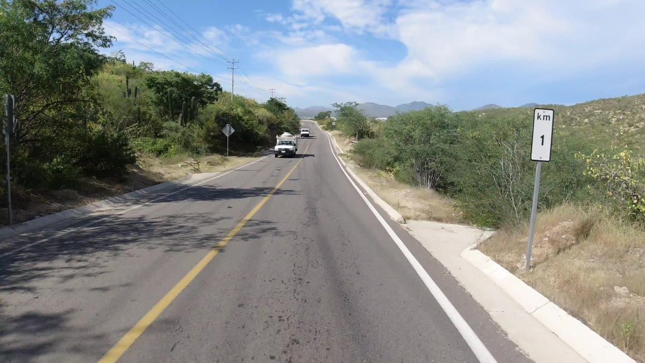 Forward view down a Los Cabos road with a distant white pickup, a kilometer marker on the right shoulder, green brush, low hills, and no visible people.