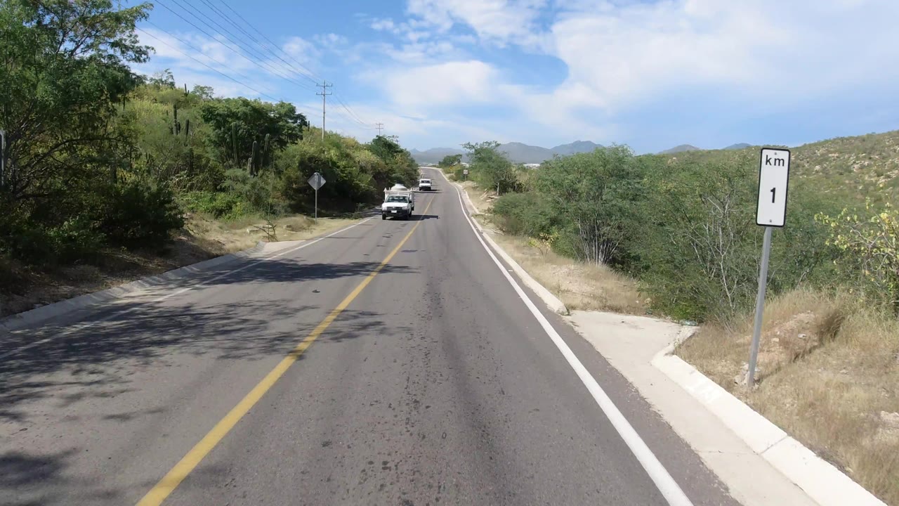 Los Cabos road view one second later, with the white pickup still ahead, the right-side kilometer marker closer to frame edge, roadside brush, hills, and no visible people.