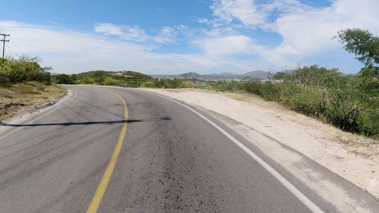 A Los Cabos road curves left past pale dirt shoulders, roadside brush, a utility pole, distant hills, blue sky, and no visible people.