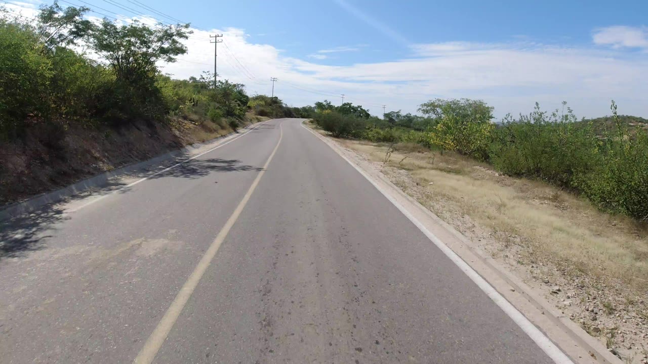 A paved two-lane road in Los Cabos, Mexico runs ahead between dry roadside vegetation, utility poles, and a blue sky, with no visible people.