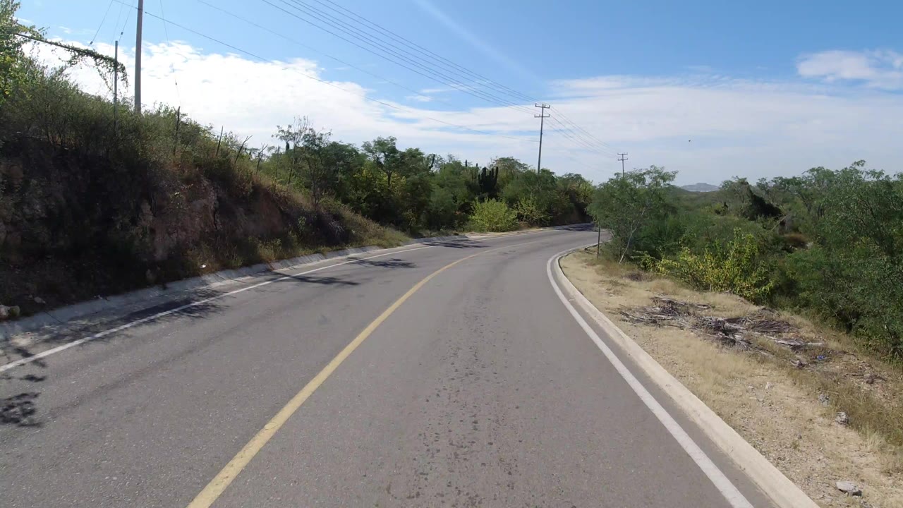 Forward road-level view on a curving Los Cabos, Mexico road with a yellow centerline, brushy shoulders, power lines, distant hills, and no visible people.
