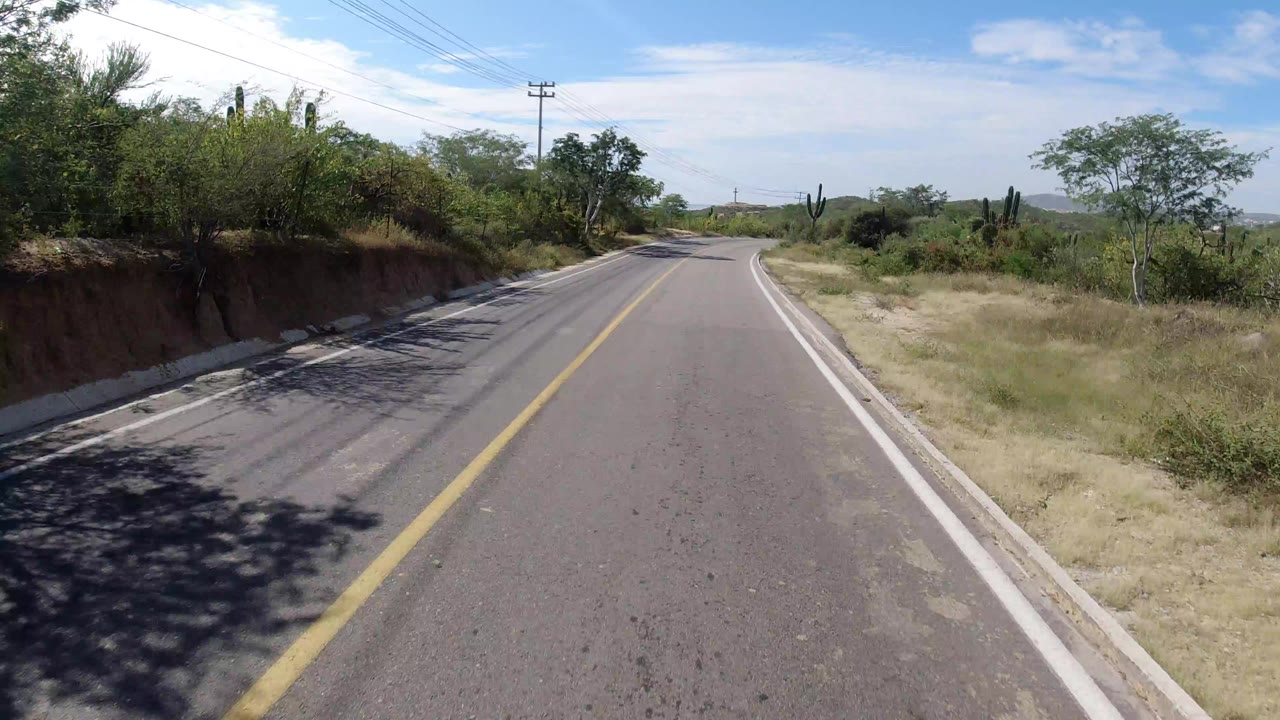 Forward road-level view in Los Cabos, Mexico, with a narrow paved road, yellow center line, brush on both sides, utility lines above, distant low hills, and no visible people.