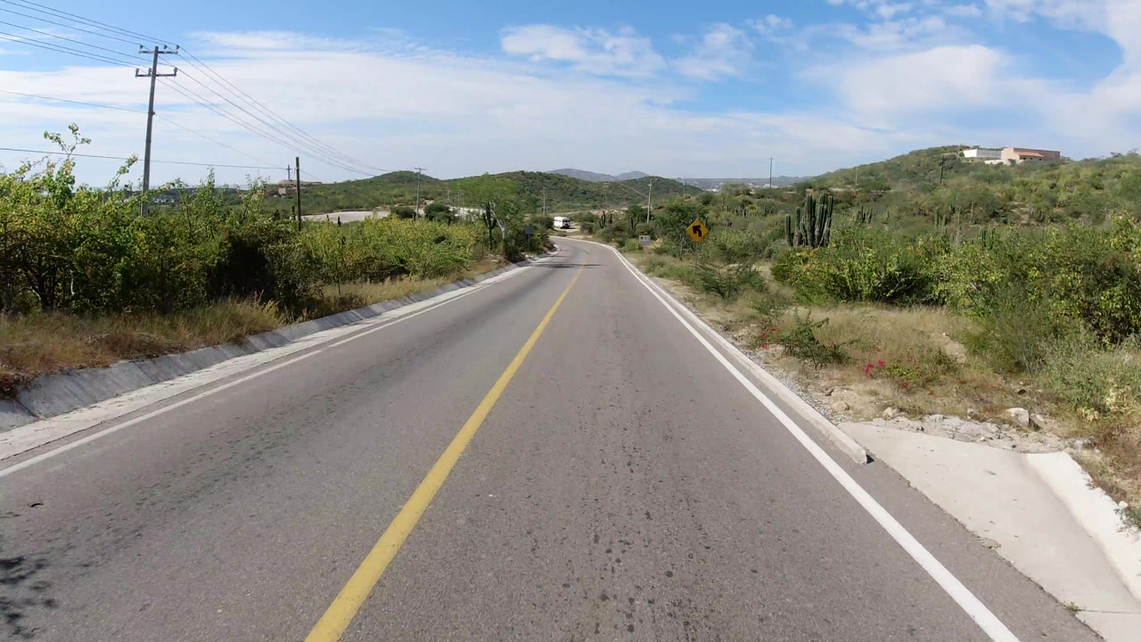 A centered road view in Los Cabos shows the curve ahead, roadside brush, overhead utility wires, a yellow curve sign, hills, and no visible people.