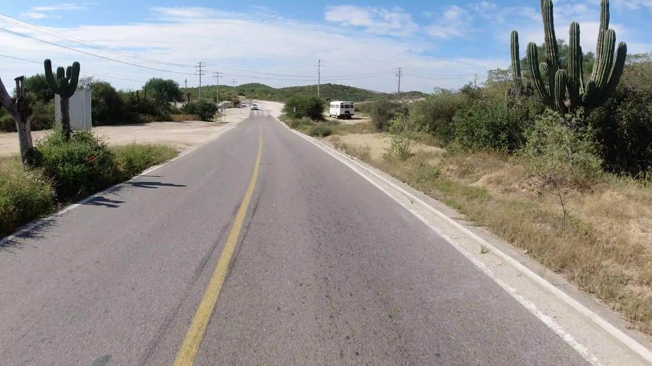 Los Cabos roadway continues past large cacti and scrub, with a white van parked off the right shoulder and no visible people.