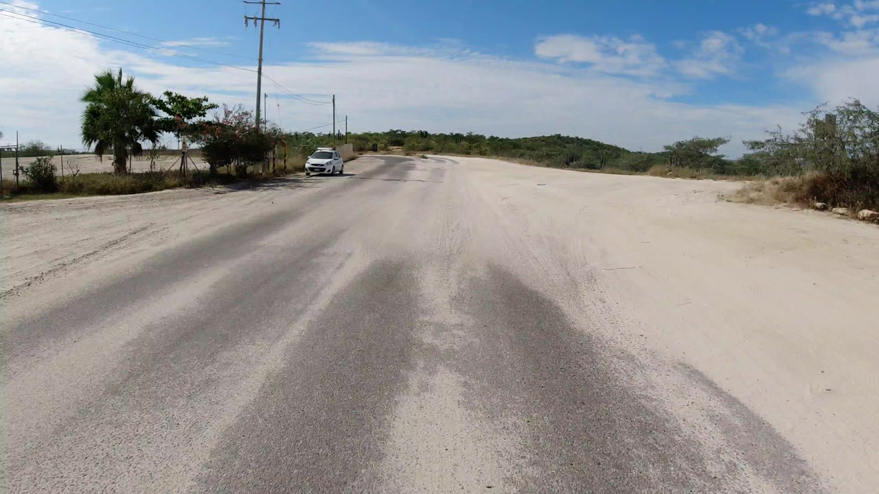 Sand-covered road with a white car, utility poles, shrubs, and blue sky.
