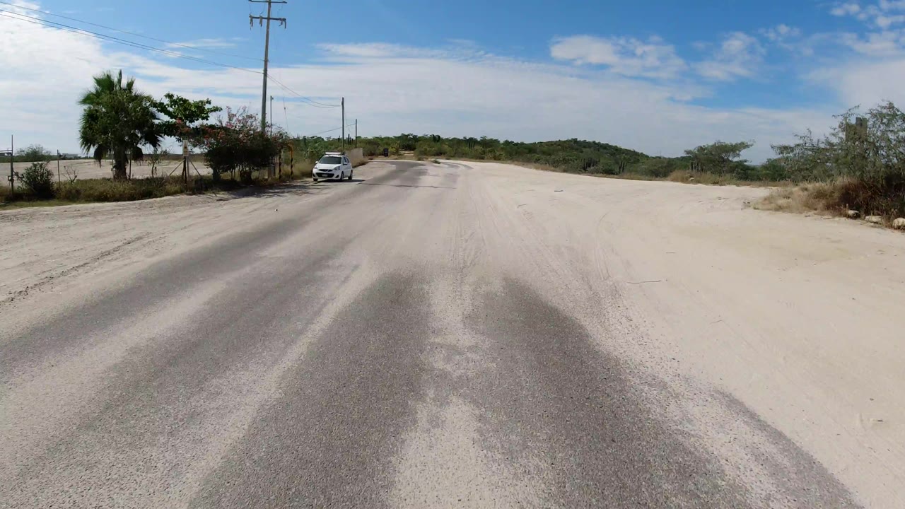 Second view in Los Cabos, Mexico, with the road stretching ahead, a white car set back on the left, roadside vegetation, utility poles, and no visible people.