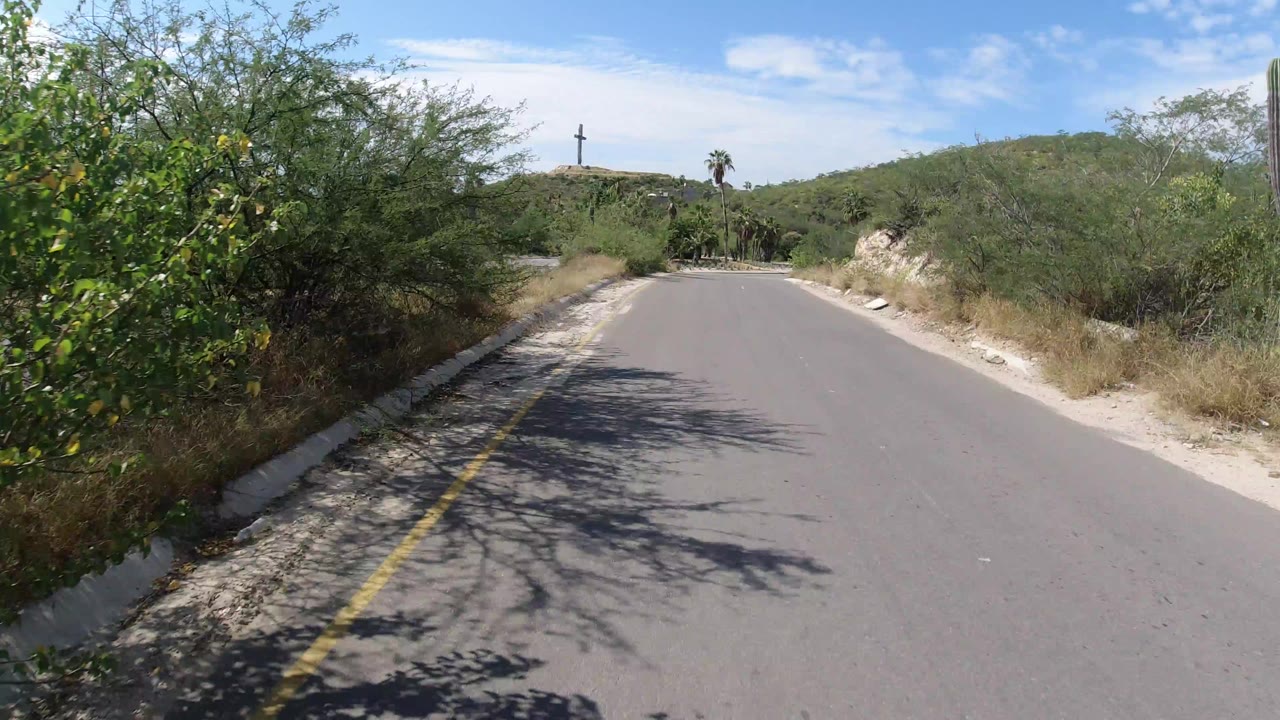 Empty paved road bordered by desert brush and a distant hilltop cross.