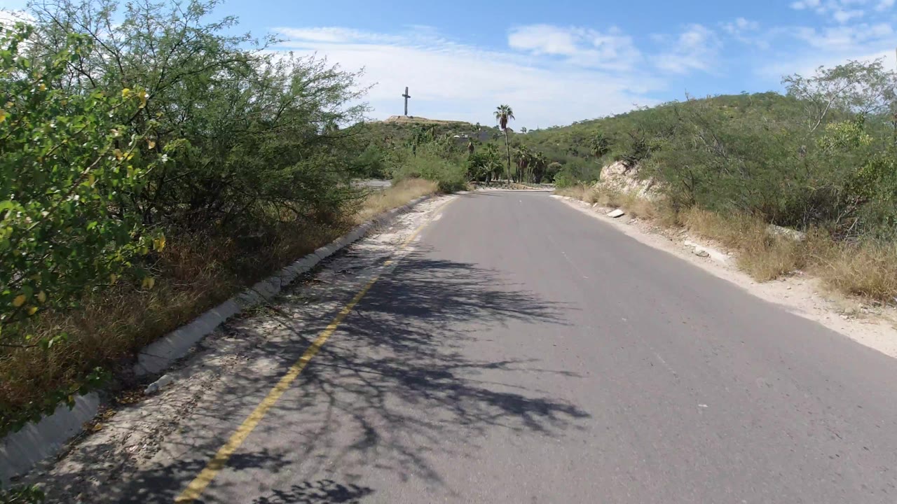 Los Cabos road frame with the lane opening wider, brush and dry grasses along both sides, pale curb stones, low hills in the distance, and no visible people.