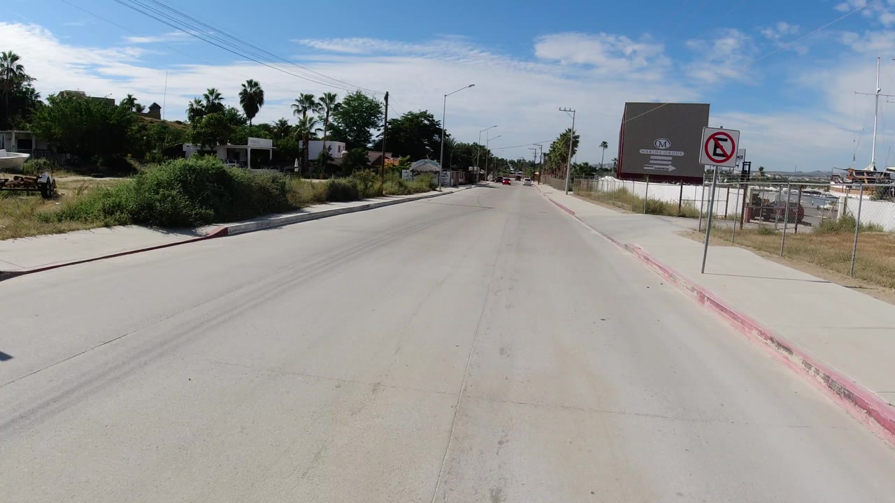 Forward street view in Los Cabos, Mexico, with a large gray commercial building on the right, planted palms on the left, overhead wires, and no clearly visible people.