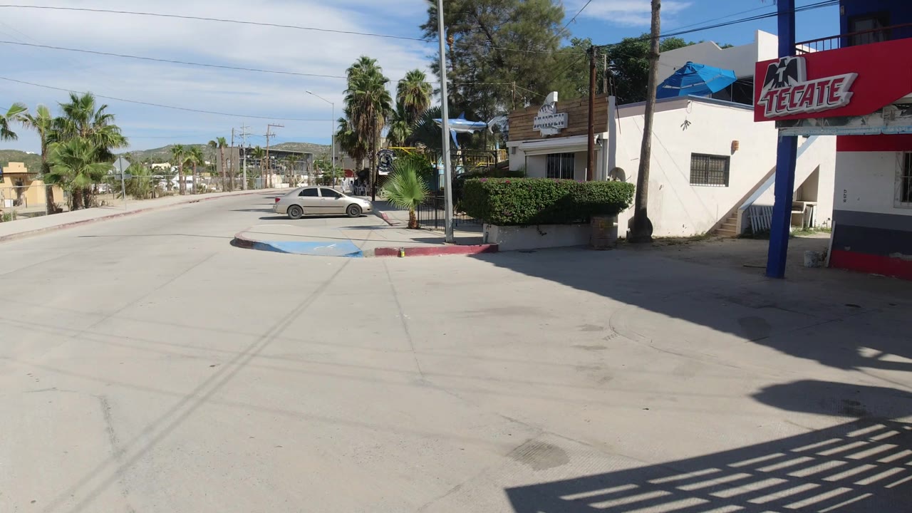 A wide Los Cabos street view shows the Tecate sign beside a low building, a parked white sedan, palms, red curbs, utility lines, and no visible people.