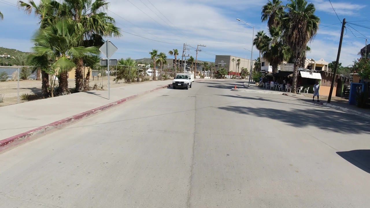 White pickup approaches on a sunlit Los Cabos street with palms, road cones, utility lines, and a shaded building frontage to the right.