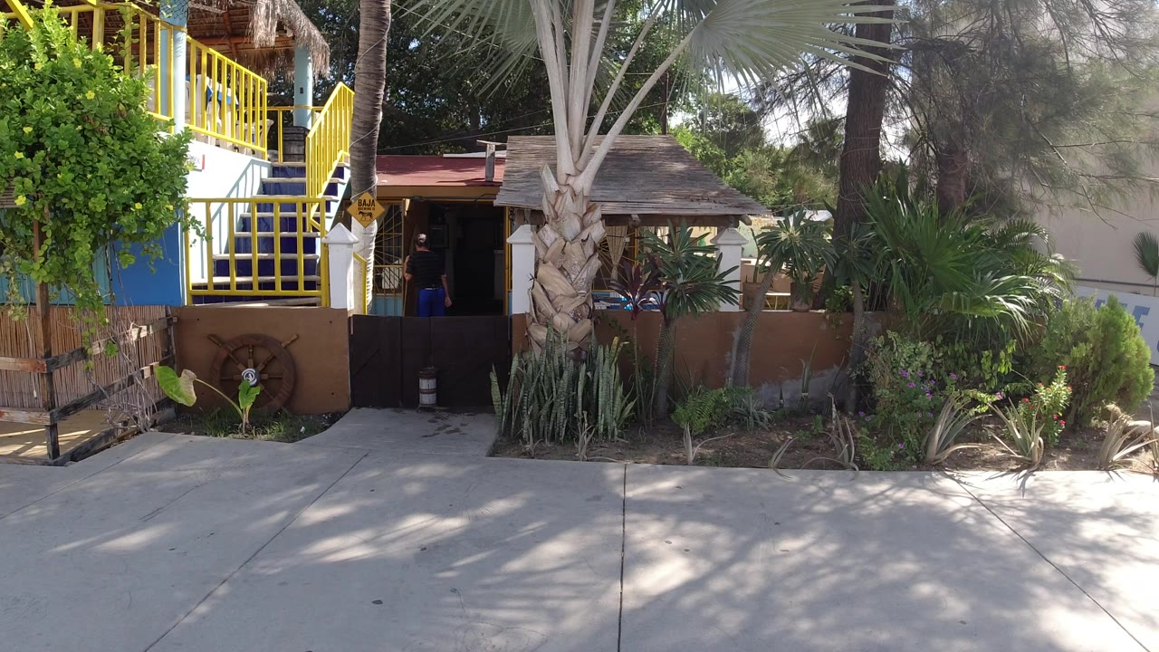 Los Cabos exterior view of the same walkway and yellow-railed stairs, with sun across the pavement and a dark doorway partly hiding a person near the entrance.