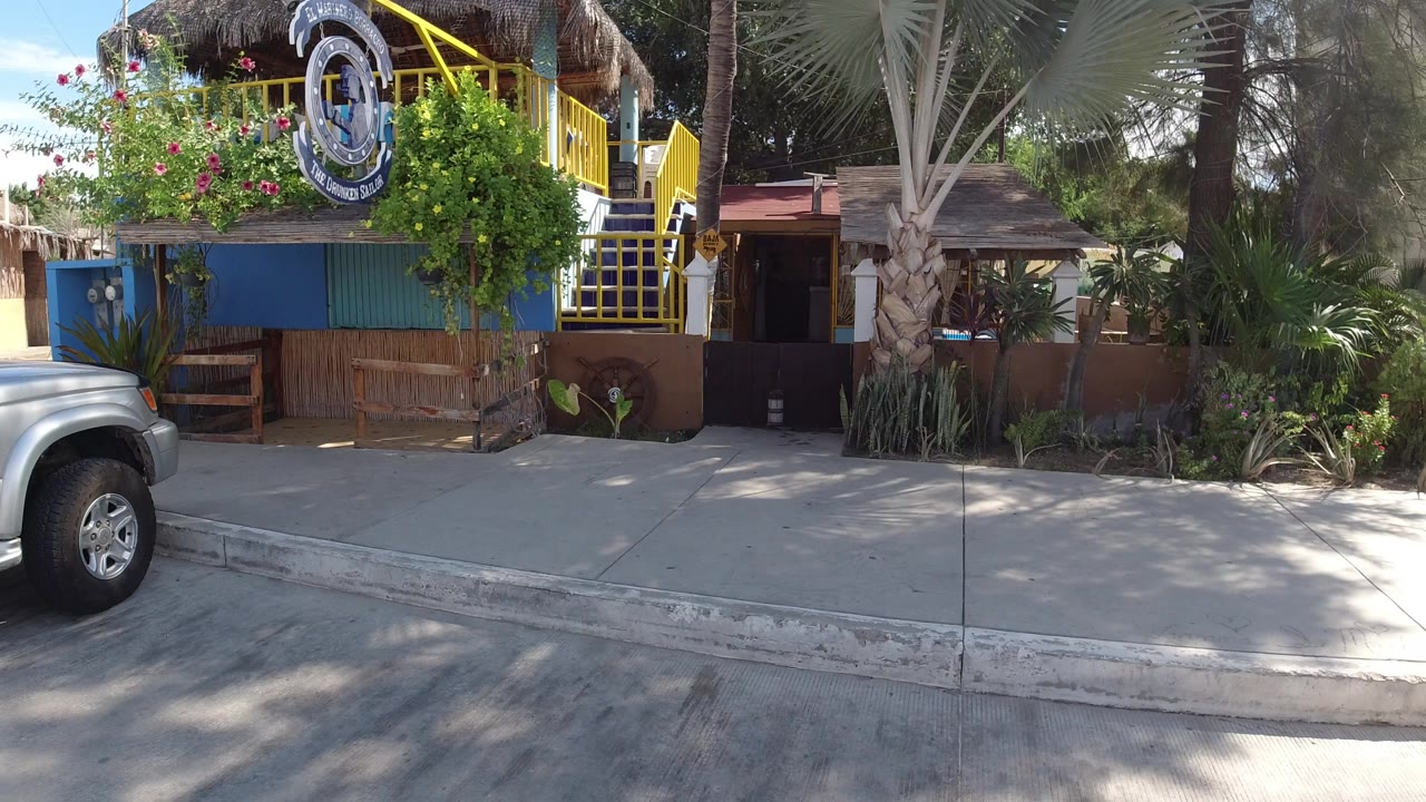 Los Cabos street-level view of a colorful house exterior with yellow railings, palm landscaping, curbside pavement, and part of a silver SUV at left, with no visible people.